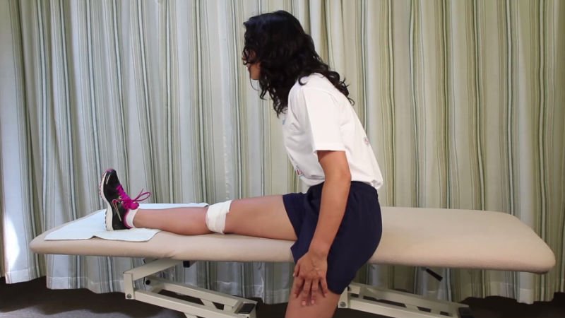 Photograph of patient seated on a plinth with the right leg supported in extension and leaning forwards.
