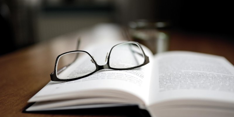Photo of a library setting, with the reader's spectacles on an open page.